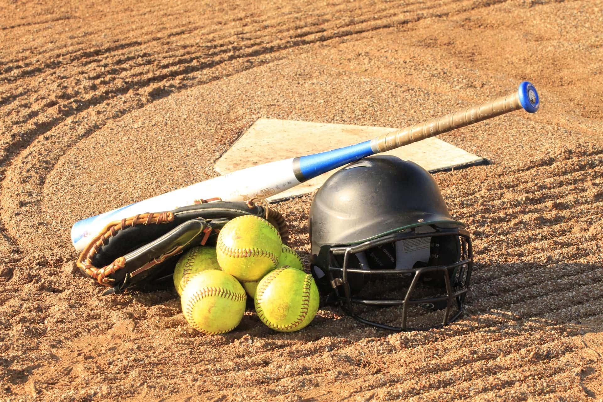 Baseball equipment including helmet, ball, bat, and glove on sandy infield, ideal for softball and baseball games.