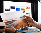 Image of person holding printed landscape photo with computer monitor displaying colorful photos, creative work.