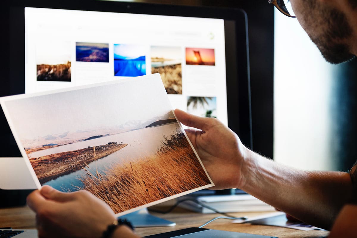 Image of person holding printed landscape photo with computer monitor displaying colorful photos, creative work.