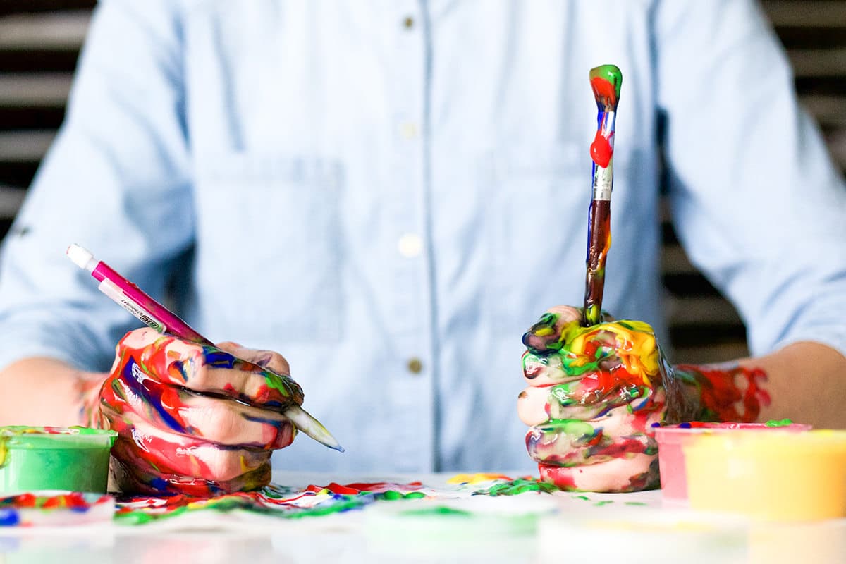 Vibrant paint-stained hands of an artist with brushes, showcasing creative process at Hoke Designs.