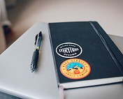Black notebook with motivational stickers, including "Design Makes Everything Possible" and travel-themed art, on a white desk.