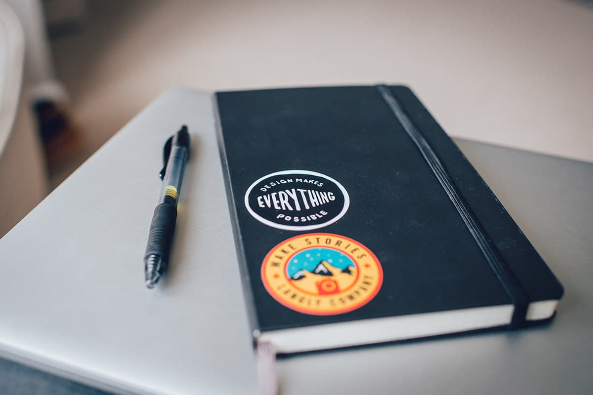 Black notebook with motivational stickers, including "Design Makes Everything Possible" and travel-themed art, on a white desk.