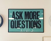 Bright blue sign with bold black text "Ask More Questions" for office or educational decor.