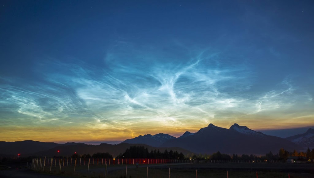 Glowing blue and white wispy clouds in a twilight sky above silhouetted mountains and a fenced field.