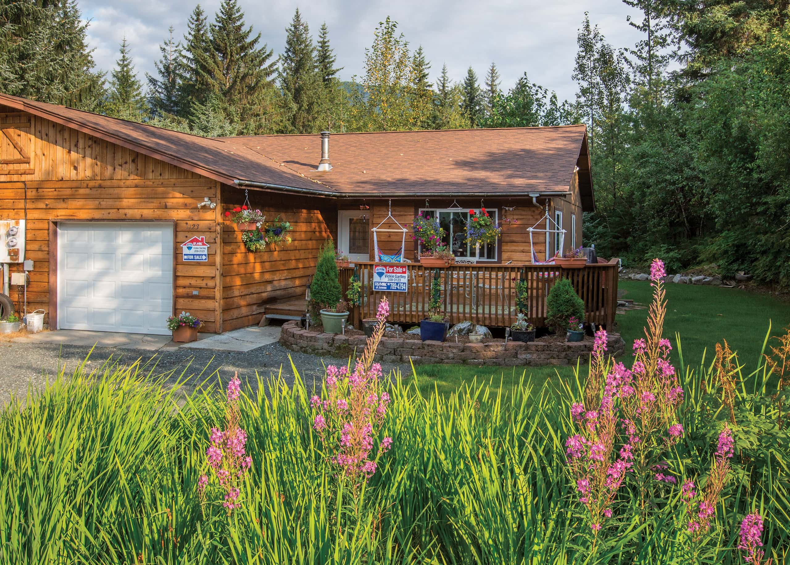 Charming wooden house with a decorated front porch and "For Sale" signs. Surrounded by lush greenery and colorful flowers.
