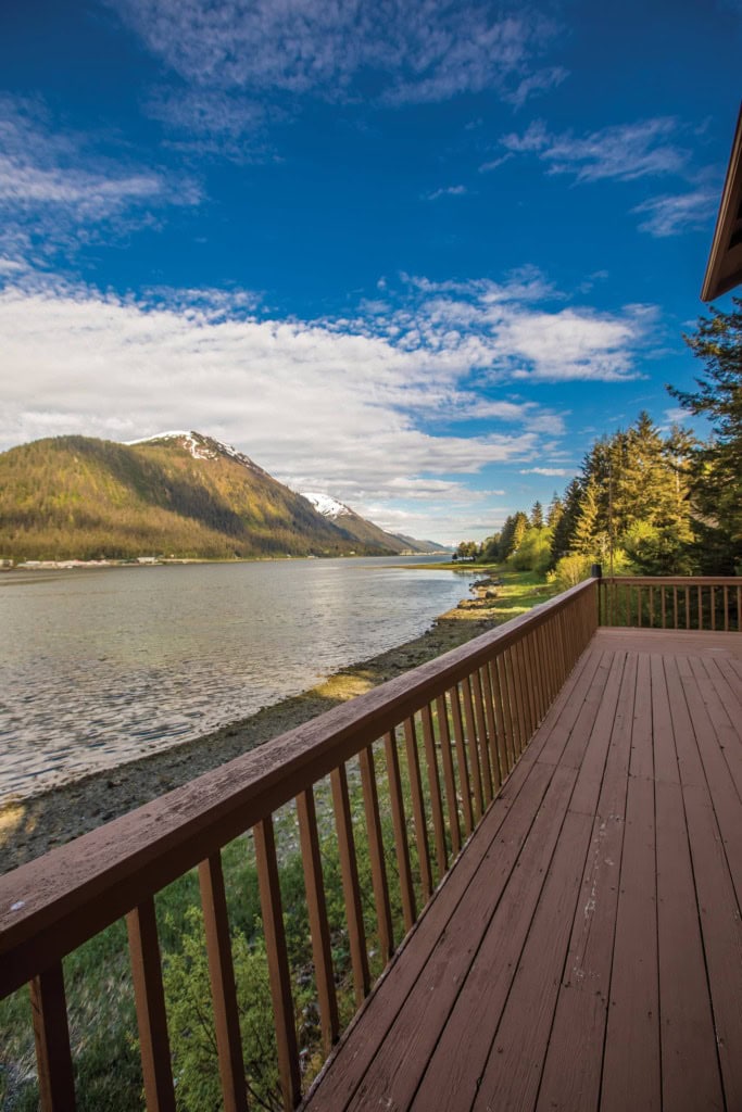 Wooden deck railing running along a rocky shoreline toward forested mountains under a blue sky with clouds.