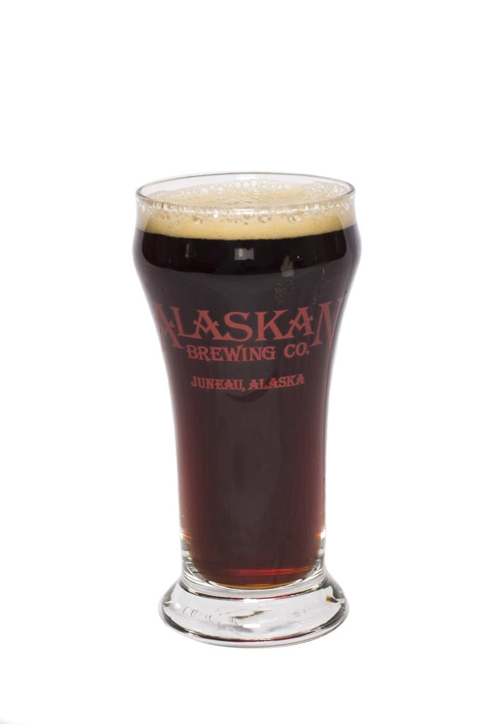Footed glass of dark amber beer printed with Alaskan Brewing Co., Juneau, Alaska in red, white background.