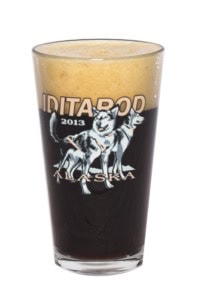 Pint glass printed with Iditarod 2013 Alaska and two sled dogs, filled with dark beer, white background.