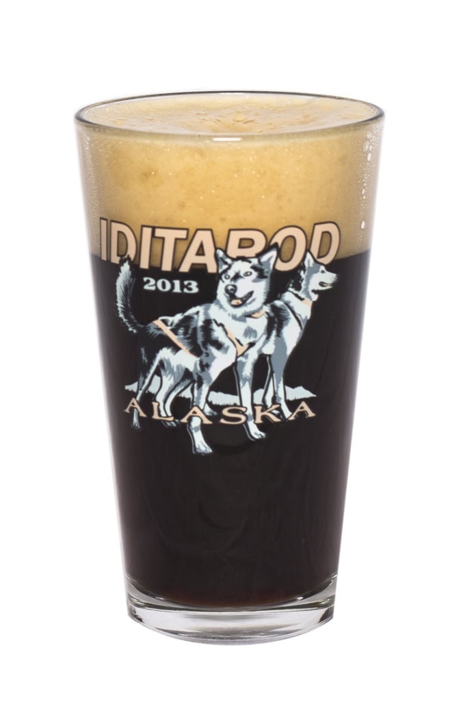 Pint glass printed with Iditarod 2013 Alaska and two sled dogs, filled with dark beer, white background.