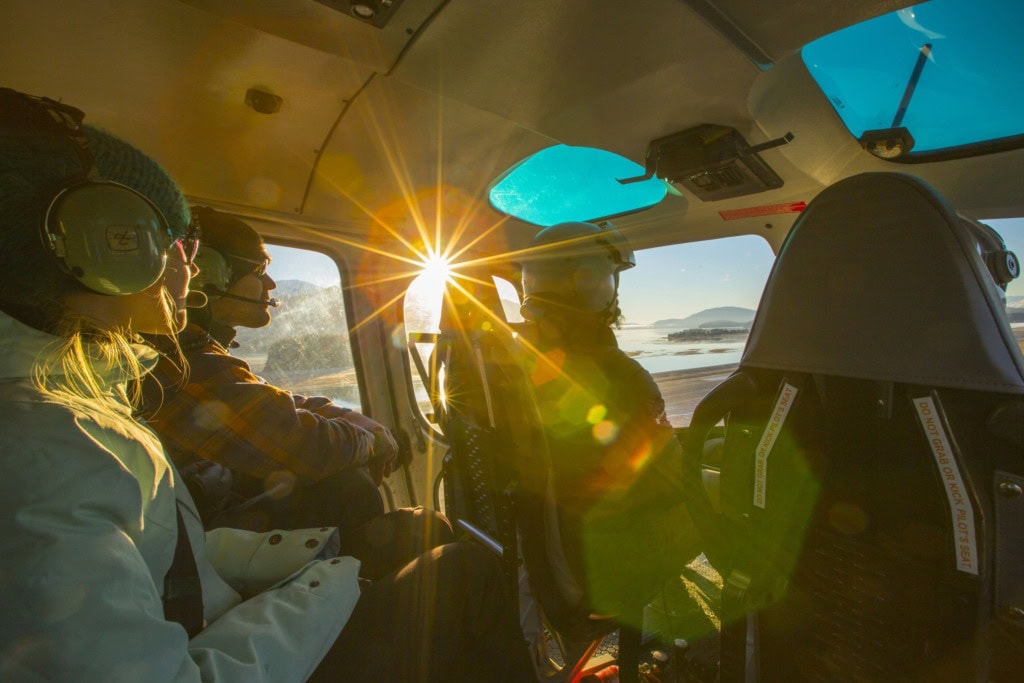 Inside a helicopter, two passengers in headsets look out the windows as sunlight flares across the cabin.