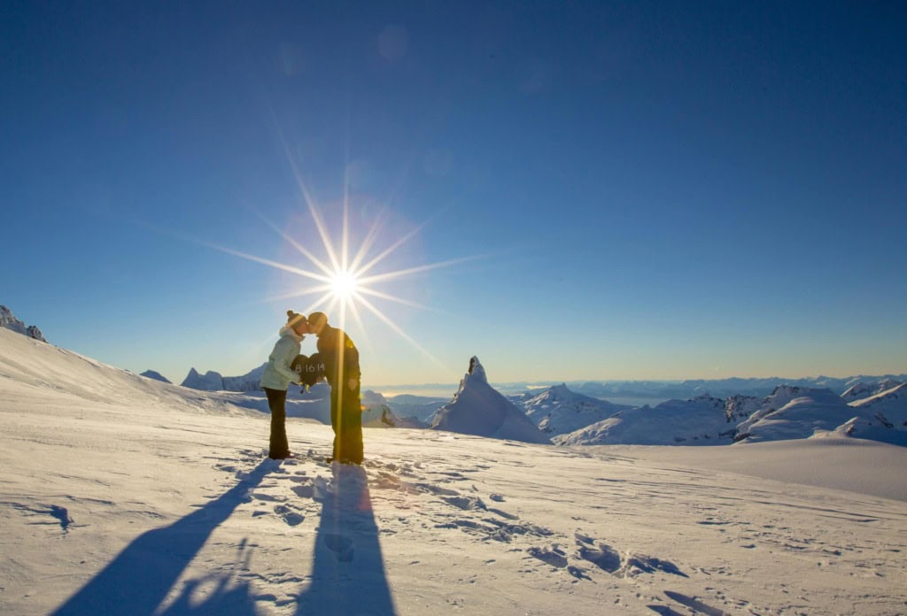 Couple kissing on a snowfield with the sun bursting behind them and snowy peaks stretching to the horizon.
