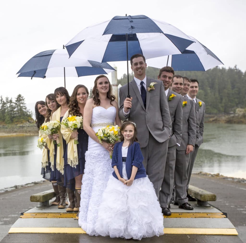Bride, groom, bridesmaids, groomsmen and a flower girl on a dock under large blue and white umbrellas.