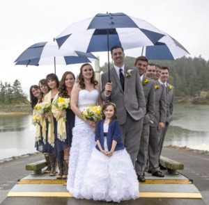 Bride, groom, bridesmaids, groomsmen and a flower girl on a dock under large blue and white umbrellas.