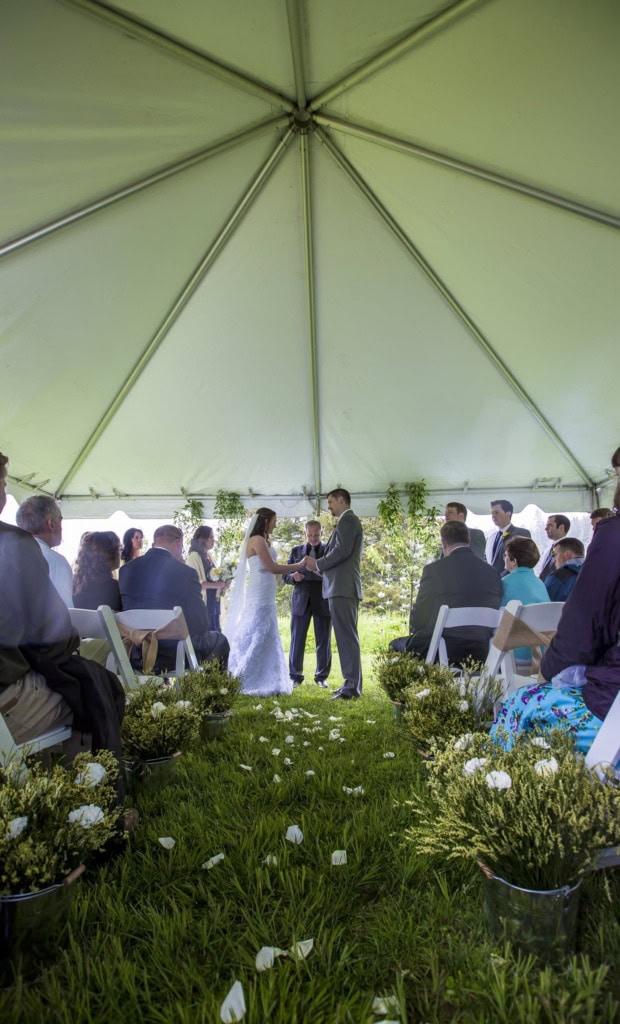 View down a grass aisle with white petals under a tent toward a bride and groom at the altar with guests seated.