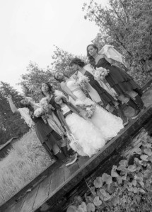 Black and white tilted photo of a bride, bridesmaids and a flower girl posing playfully on a wooden boardwalk.