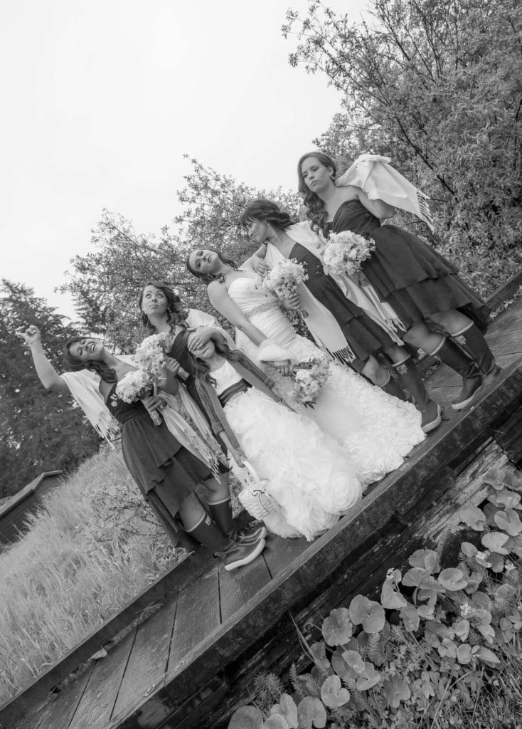 Black and white tilted photo of a bride, bridesmaids and a flower girl posing playfully on a wooden boardwalk.
