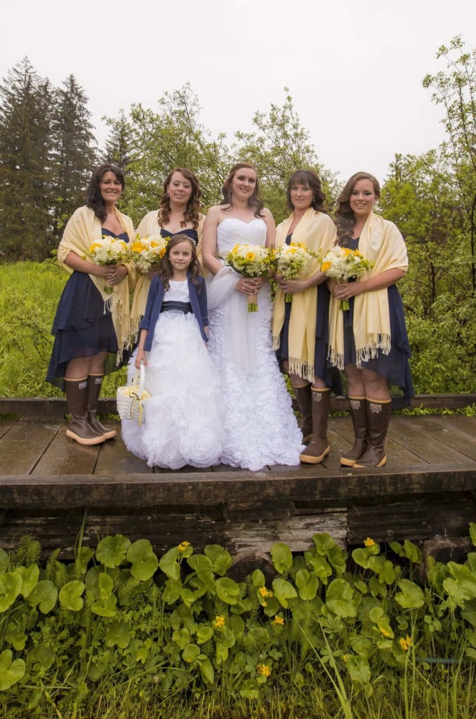 Bride with four bridesmaids in navy dresses, yellow shawls and rubber boots, plus a flower girl, on a boardwalk.