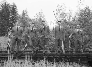 Black and white photo of five groomsmen in suits standing on a wooden platform holding striped umbrellas.