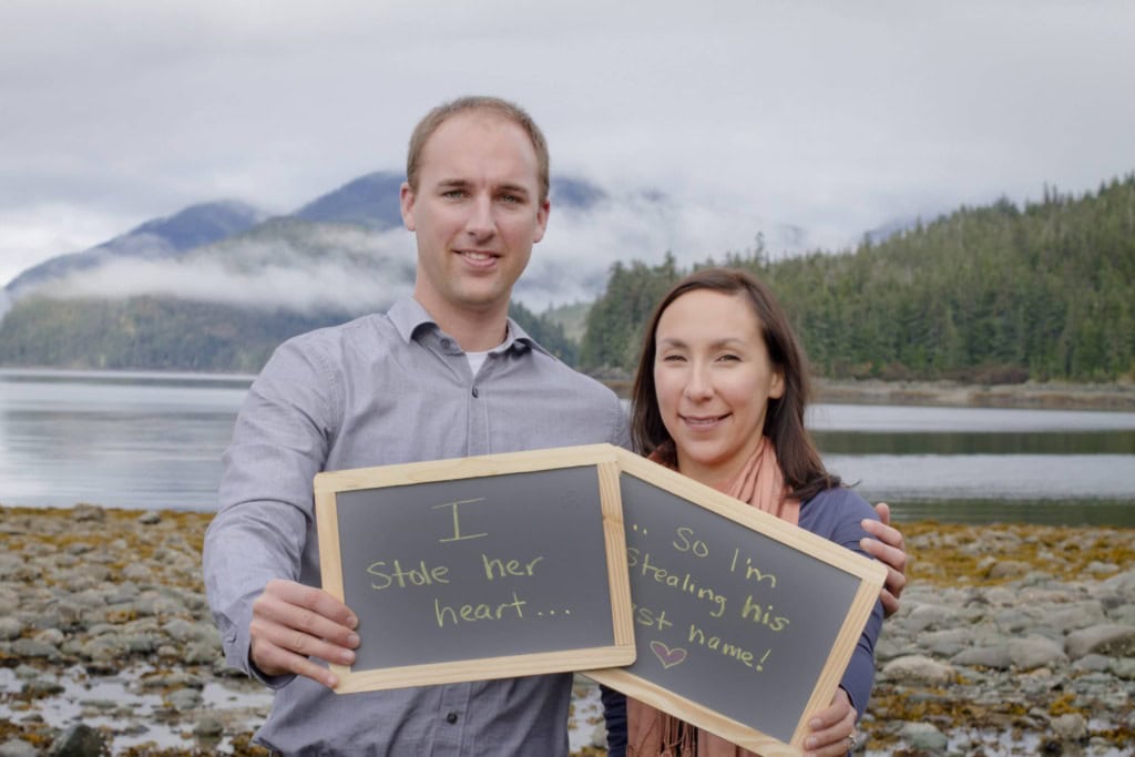 Couple holding chalkboards reading I stole her heart, so I'm stealing his last name, on a rocky beach.