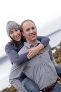 Smiling woman in a knit hat riding piggyback on a man in a gray shirt, with water and rocks behind.