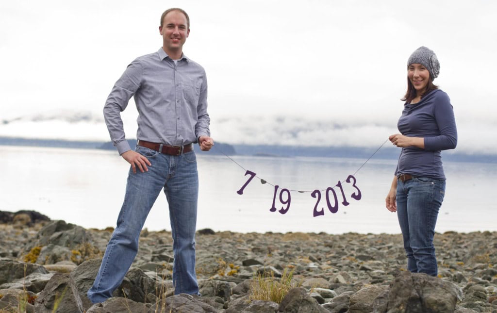 Couple on a rocky beach holding a string banner reading 7 19 2013, with water and low clouds behind.