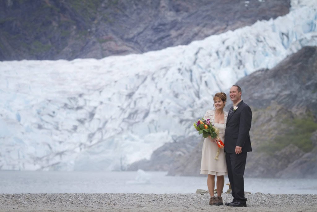 Couple in wedding attire on a gravel beach, smiling, with a large glacier and mountain rising behind them.
