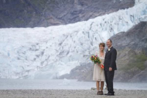 Couple in wedding attire on a gravel beach, smiling, with a large glacier and mountain rising behind them.