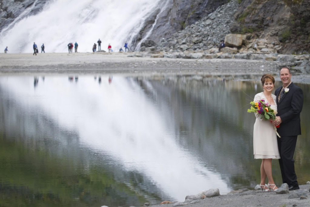 Couple in wedding attire at the edge of a still lake reflecting a large waterfall, with visitors in the distance.