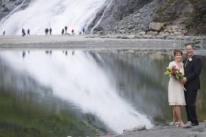Couple in wedding attire at the edge of a still lake reflecting a large waterfall, with visitors in the distance.