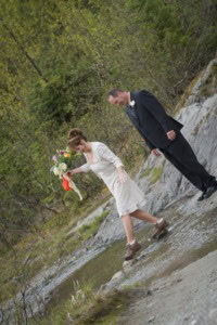 Bride in hiking boots steps across a shallow creek holding a bouquet while the groom leans to watch.