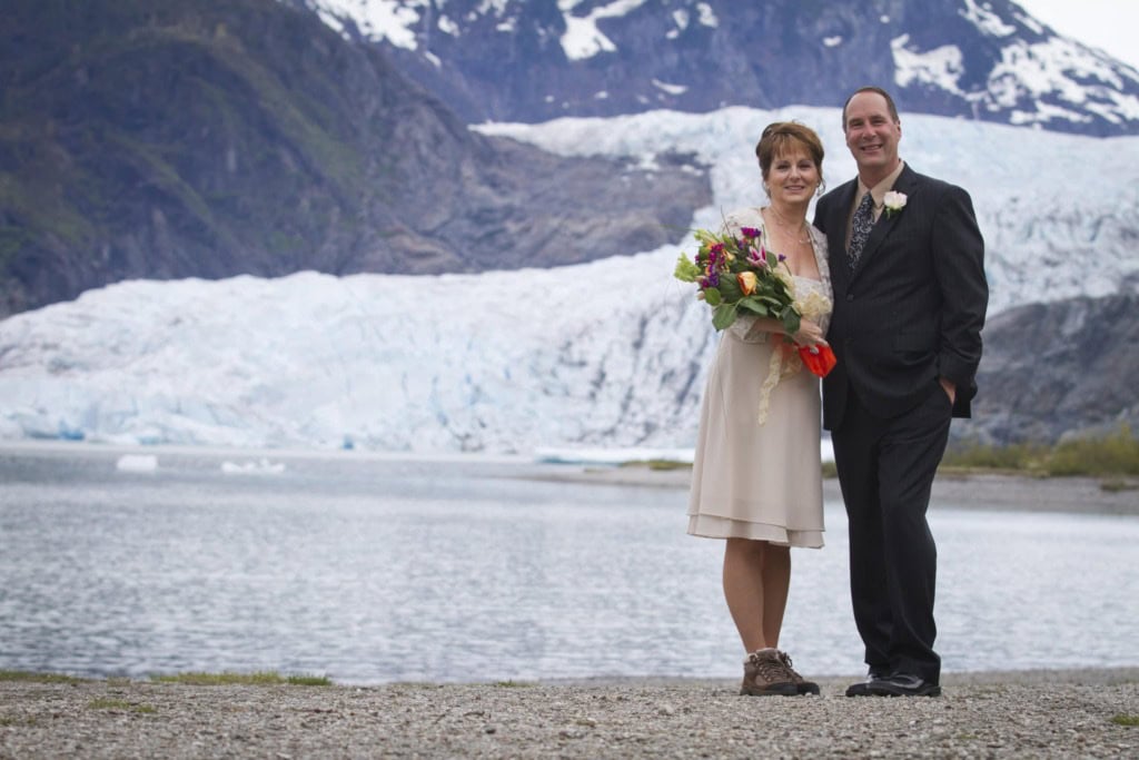 Couple in wedding attire standing on a gravel shore in front of a lake and a glacier.