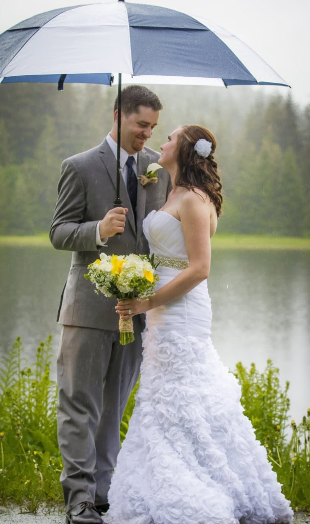 Bride and groom sharing a blue and white umbrella beside a misty lake, the bride holding a yellow and white bouquet.