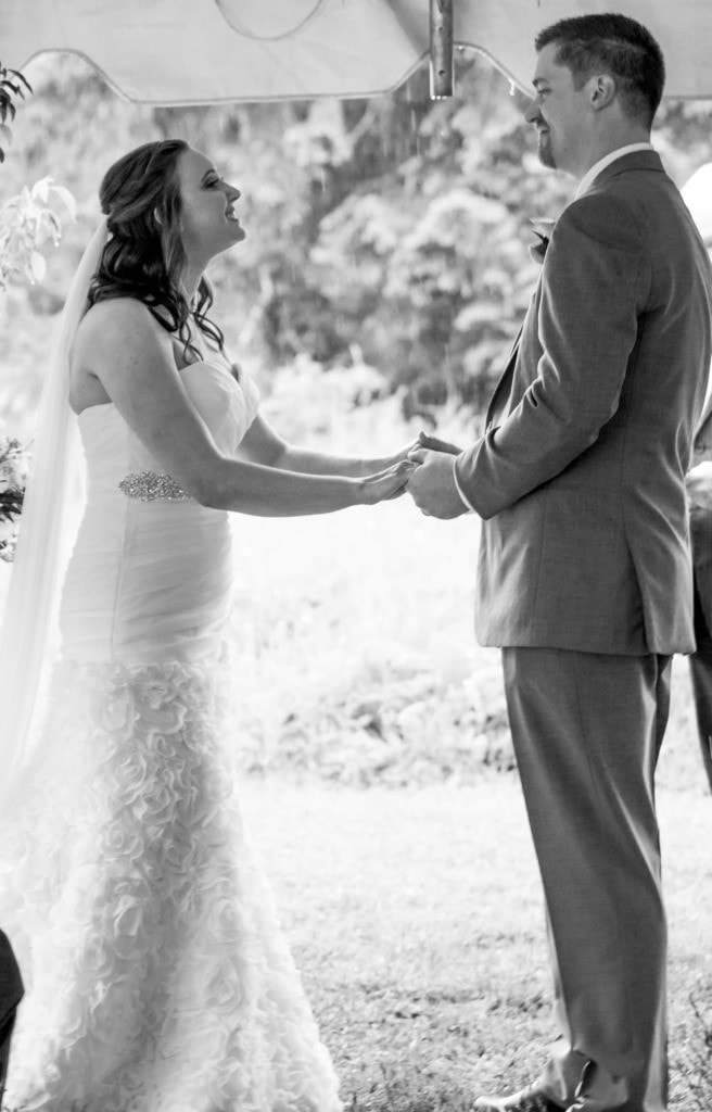 Black and white photo of a bride and groom holding hands and smiling at each other during their vows.