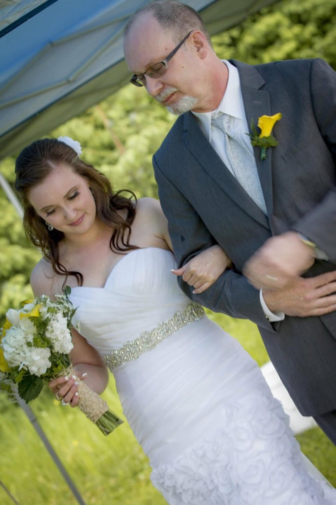 Bride in a strapless gown with a bouquet walks arm in arm with an older man in a gray suit.