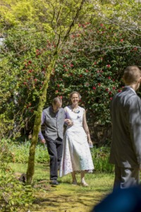 Bride in a white lace dress walks arm in arm with an older man along a garden path lined with red flowers.