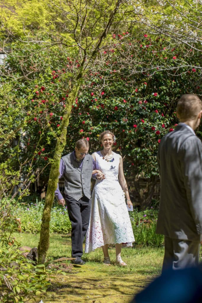 Bride in a white lace dress walks arm in arm with an older man along a garden path lined with red flowers.