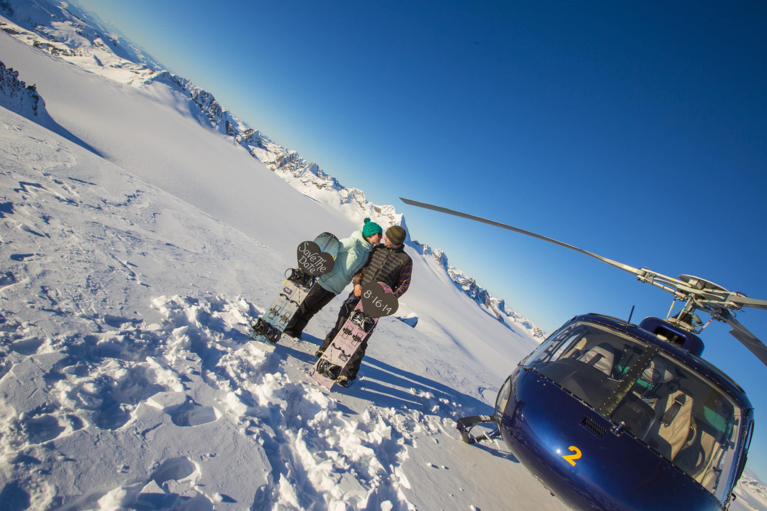 Two snowboarders sharing a kiss next to a helicopter on snowy mountain with clear blue sky.