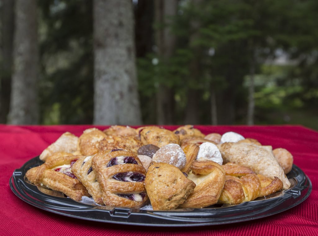 Tray of danishes, scones and powdered donut holes on a red tablecloth with tall trees behind.