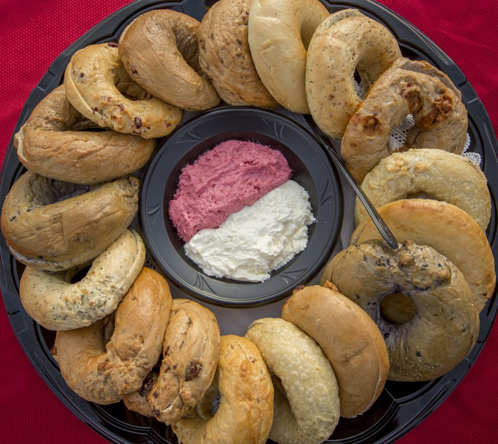 Overhead view of assorted bagels around a dish of berry and plain cream cheese on a red tablecloth.