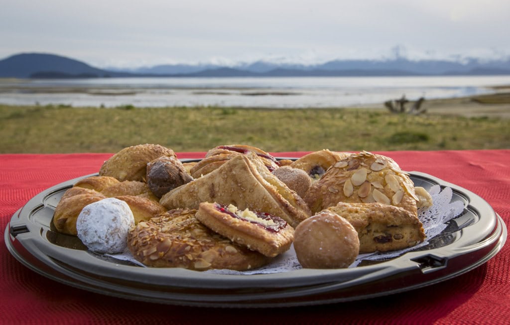 Tray of almond pastries, danishes and donut holes on a red table with grass, water and mountains behind.