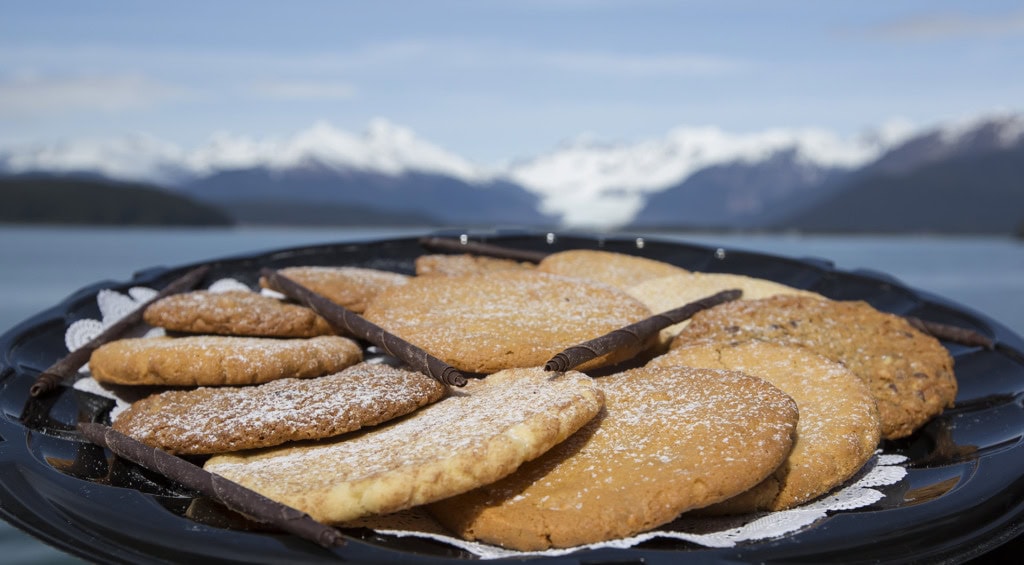 Black tray of sugar-dusted cookies with chocolate rolls, snowy mountains and water blurred behind.