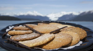 Black tray of sugar-dusted cookies with chocolate rolls, snowy mountains and water blurred behind.