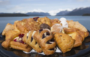 Pile of pastries with blueberry and cherry danishes, almond pastries and powdered donuts, mountains behind.