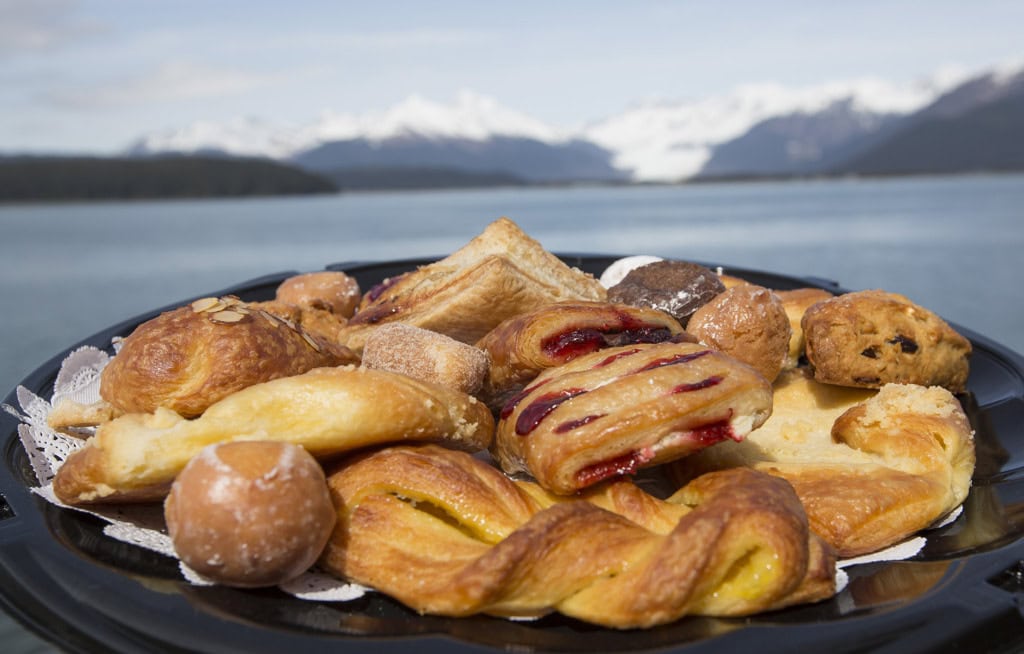 Tray of pastries including cherry danish, twists and donut holes, snowy mountains and water in the background.