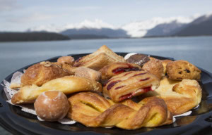 Tray of pastries including cherry danish, twists and donut holes, snowy mountains and water in the background.
