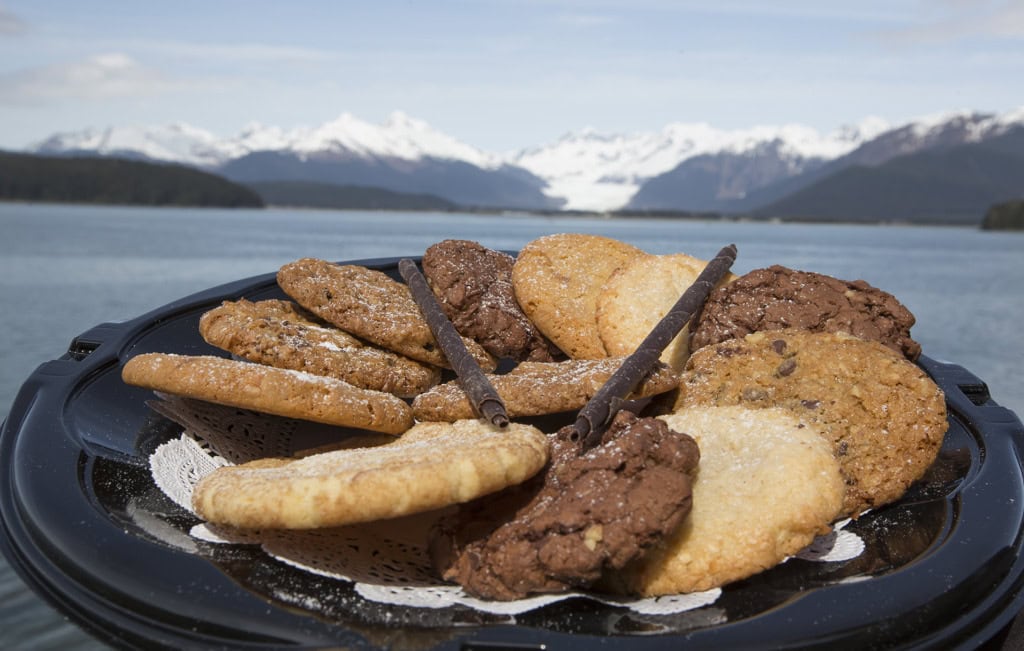 Tray of assorted cookies with chocolate rolls on a doily, snowy mountains and water behind it.