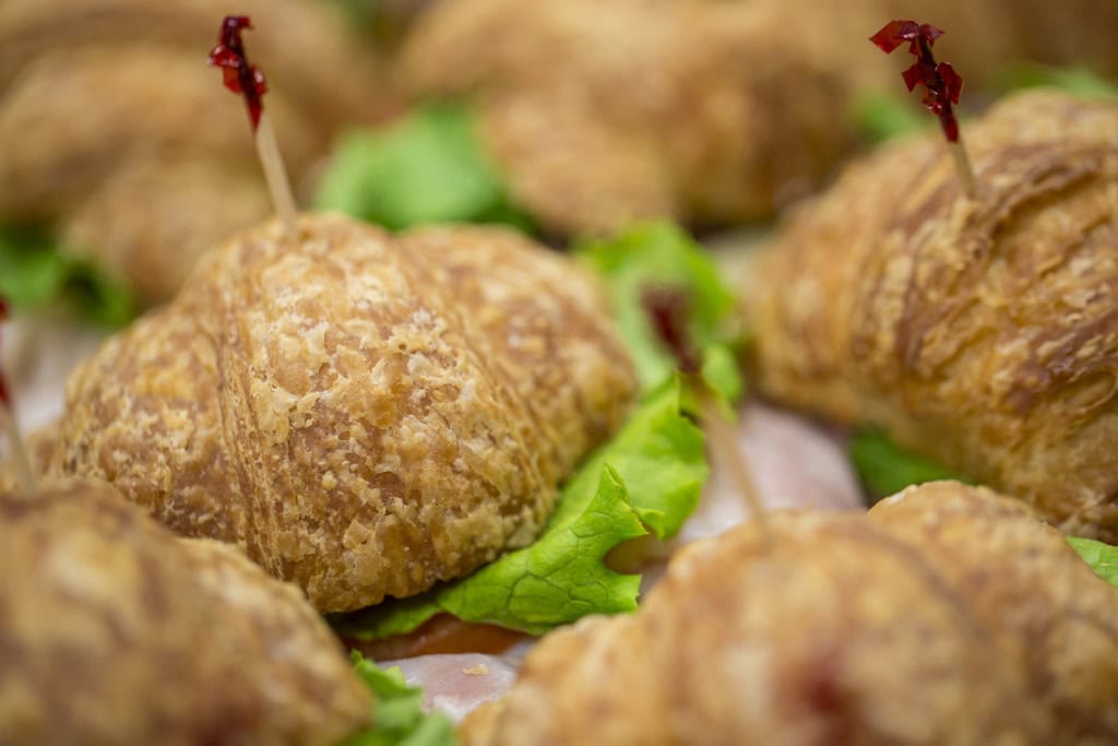 Close-up of a flaky croissant sandwich with green lettuce and a red decorative pick, others blurred around it.