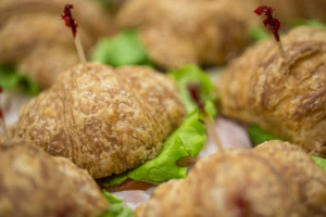 Close-up of a flaky croissant sandwich with green lettuce and a red decorative pick, others blurred around it.