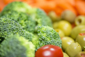 Close-up of broccoli florets, green olives and a cherry tomato with carrots blurred in the background.