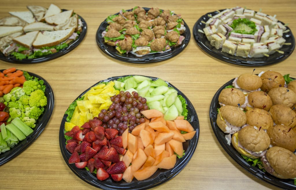 Six party platters on a wood table: sandwiches, croissant sandwiches, finger sandwiches, vegetables, fruit, sliders.